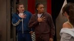 "Two surprised young men in tracksuits stand indoors with hands on chests, facing a woman in a casual outfit. The room has red curtains and appears to be part of a modern home or apartment."