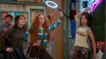 "Three stylish teens pose confidently in a colorful school hallway, one holding a ring light, with lockers, posters, and a neon sign in the background. Other students are visible further down the hall."