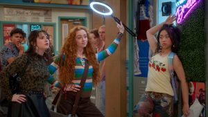 "Three stylish teens pose confidently in a colorful school hallway, one holding a ring light, with lockers, posters, and a neon sign in the background. Other students are visible further down the hall."