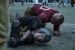 "Two vintage football players wearing old-style helmets and uniforms lie on a muddy field after a tackle, both looking exhausted; blurred players and spectators are seen in the background during a gritty, historical game."