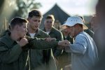 "Military recruits practice hand-to-hand combat outdoors, wearing green uniforms, with a trainer in a bucket hat instructing them; tents and trees visible in the background, group setting, training camp atmosphere."