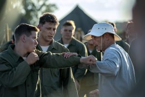 "Military recruits practice hand-to-hand combat outdoors, wearing green uniforms, with a trainer in a bucket hat instructing them; tents and trees visible in the background, group setting, training camp atmosphere."