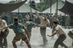 "Soldiers in vintage uniforms play football on a dusty field surrounded by military tents, with other soldiers watching, suggesting a break in a wartime or military camp setting."