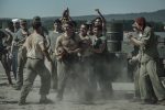"A group of WWII-era soldiers in dusty uniforms are shouting and cheering around two men, possibly during a brawl or contest, outdoors near metal barrels and mountains in the background."