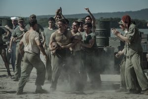 "A group of WWII-era soldiers in dusty uniforms are shouting and cheering around two men, possibly during a brawl or contest, outdoors near metal barrels and mountains in the background."