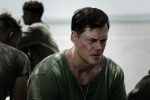 "Bill Skarsgard as Bob Baumann in a military-style green shirt sits outdoors near water, looking serious and thoughtful, with other men in similar attire blurred in the background suggesting a somber, reflective scene."