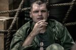 "Ray Nicholson as Tony Butkovich in green fatigues sits against wooden crates and ropes, holding a cigarette. He wears dog tags and has a serious expression, suggesting a tense or contemplative moment in a military setting."