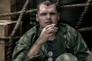 "Ray Nicholson as Tony Butkovich in green fatigues sits against wooden crates and ropes, holding a cigarette. He wears dog tags and has a serious expression, suggesting a tense or contemplative moment in a military setting."