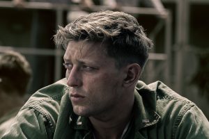"Tom Francis as Dave Schreiner in military uniform with short hair looks to the side with a serious expression, outdoors with blurred structures and another person in the background, suggesting a wartime or military setting."