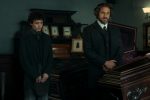 "Two men in dark, old-fashioned suits stand beside a polished wooden coffin in a dimly lit, ornate room decorated with framed photos, suggesting a 19th-century funeral or wake setting."