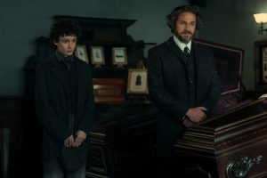 "Two men in dark, old-fashioned suits stand beside a polished wooden coffin in a dimly lit, ornate room decorated with framed photos, suggesting a 19th-century funeral or wake setting."