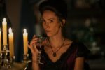 "Woman in period costume sits at a candlelit table, holding a glass of red wine, with a serious expression in a dimly lit, elegant room."