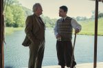 "Two men in aristocratic clothing talk by a riverbank; one holds a drink, the other a walking stick. They stand under a shaded structure with green trees and calm water visible in the background on a sunny day."
