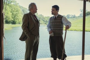 "Two men in aristocratic clothing talk by a riverbank; one holds a drink, the other a walking stick. They stand under a shaded structure with green trees and calm water visible in the background on a sunny day."