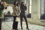 "Two stylish women stand in an elegant, classic interior with tall windows and ornate decor. One holds a white handbag, both wear blazers and boots, appearing poised and fashionable in a well-lit, sophisticated room with flowers and art."