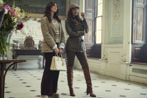 "Two stylish women stand in an elegant, classic interior with tall windows and ornate decor. One holds a white handbag, both wear blazers and boots, appearing poised and fashionable in a well-lit, sophisticated room with flowers and art."