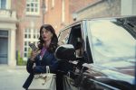 "Woman with long dark hair in a stylish blazer stands next to a black luxury car on a city street, holding a smartphone and beige purse, with upscale buildings in the background."
