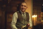 "Middle-aged man in a checkered shirt and vest holding a book, standing indoors with warm lighting, blurred background features candles and wooden decor, creating a cozy, intimate atmosphere."
