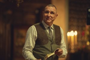 "Middle-aged man in a checkered shirt and vest holding a book, standing indoors with warm lighting, blurred background features candles and wooden decor, creating a cozy, intimate atmosphere."