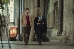 "Two well-dressed men walking together inside a grand, historic stone building with high ceilings and stained glass windows, suggesting a church or cathedral setting."