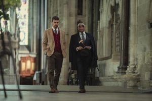 "Two well-dressed men walking together inside a grand, historic stone building with high ceilings and stained glass windows, suggesting a church or cathedral setting."