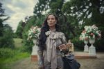 "Woman with long dark hair in an elegant outfit and bow scarf stands outdoors in a lush garden with large floral arrangements and greenery, holding a black handbag on her arm."