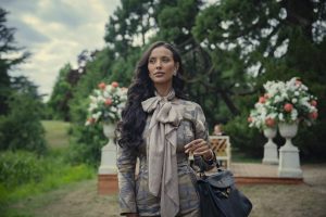 "Woman with long dark hair in an elegant outfit and bow scarf stands outdoors in a lush garden with large floral arrangements and greenery, holding a black handbag on her arm."