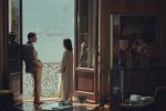 "A man and woman stand on a balcony overlooking a lake, seen from inside a richly decorated room with large open doors, elegant furniture, a vase of flowers, and mountains visible in the background."