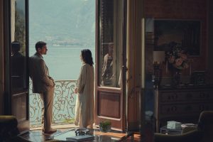 "A man and woman stand on a balcony overlooking a lake, seen from inside a richly decorated room with large open doors, elegant furniture, a vase of flowers, and mountains visible in the background."