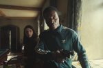 "A man in a green tracksuit holds a gun in a dimly lit room, appearing tense, while a woman stands behind him looking concerned; patterned curtains and home interior details are visible."