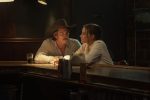 "A man in a cowboy hat and a woman sit closely at a dimly lit bar, talking seriously. Glasses of drink rest on the bar, and the setting is cozy with warm lighting and rustic wood and brick surroundings."