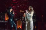 "Two singers performing on stage, one in a black outfit with a hat and the other in a white dress, with a live band playing instruments in the background and dramatic stage lighting."