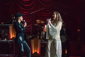 "Two singers performing on stage, one in a black outfit with a hat and the other in a white dress, with a live band playing instruments in the background and dramatic stage lighting."