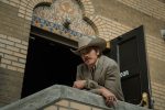 "Man in a cowboy hat and suit leans on a concrete railing outside a brick building with decorative arch, open door and ornate tilework above. Daylight scene, western style clothing, casual pose."