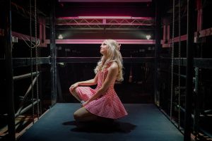 "Woman in a pink checkered dress kneeling on a stage or backstage area, with dramatic lighting and industrial surroundings, looking upward under pink overhead lights."