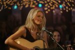 "A woman with long blonde hair plays an acoustic guitar and sings into a microphone on stage under string lights, surrounded by an audience in a dimly lit, cozy music venue."