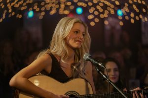 "A woman with long blonde hair plays an acoustic guitar and sings into a microphone on stage under string lights, surrounded by an audience in a dimly lit, cozy music venue."