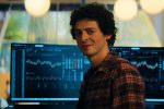 "A man in a plaid shirt sits in front of computer monitors displaying financial charts and stock data in a modern office with large windows and warm lighting."