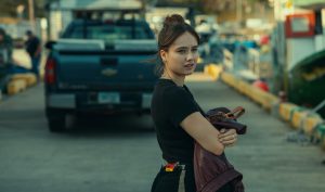 "Young woman holding a brown jacket stands on a dock near parked trucks, looking back with a serious expression, in an outdoor marina environment with boats and industrial equipment in the background."