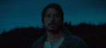 "A man with facial hair stands outdoors at dusk, looking ahead with a concerned expression, silhouetted trees and a dark blue evening sky in the background."