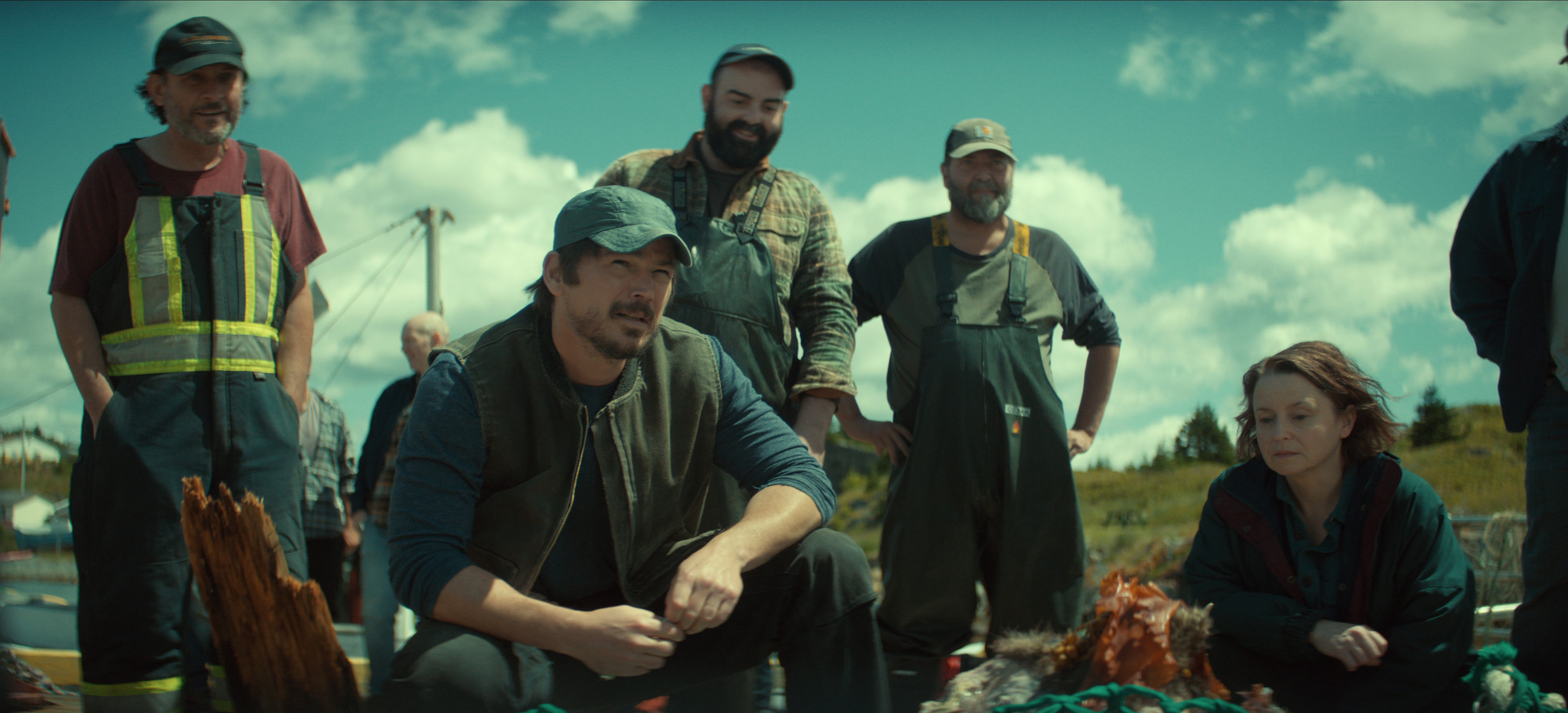 "A group of fishermen and a woman crouch and stand outdoors near fishing gear under a bright blue sky with clouds, on a grassy landscape possibly near the sea."