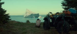 "Person sitting on grass near an ATV, overlooking a large iceberg in the ocean, framed by trees on both sides, under a clear sky."