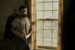 "A man with a beard stands pensively near a sunlit window in a rustic room, dressed in an old-fashioned shirt and trousers. The environment suggests a historical or rural setting with muted colors and a calm mood."