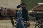 "A woman and man in period clothing walk beside a horse-drawn wagon loaded with luggage and crates, set in a rustic outdoor location, evoking a historical setting."