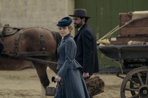 "A woman and man in period clothing walk beside a horse-drawn wagon loaded with luggage and crates, set in a rustic outdoor location, evoking a historical setting."