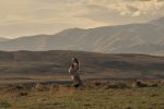 "Pregnant woman standing in a grassy field at sunset, surrounded by wildflowers, with rolling hills and mountains in the background under a cloudy sky."
