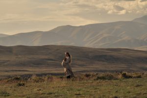 "Pregnant woman standing in a grassy field at sunset, surrounded by wildflowers, with rolling hills and mountains in the background under a cloudy sky."