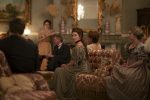 "A group of people in period clothing sit in an ornate, vintage living room with elegant furniture, antique decorations, and a large mirror above the fireplace, engaged in conversation or listening."