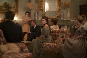 "A group of people in period clothing sit in an ornate, vintage living room with elegant furniture, antique decorations, and a large mirror above the fireplace, engaged in conversation or listening."