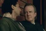"Two men sit close together against a wooden wall, one gripping the other’s neck while they exchange an intense stare, suggesting a dramatic or emotional moment in a historical or rustic indoor setting."